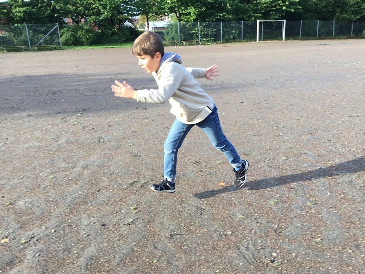 Ayham har blå byxor och ljus jacka. Han är mitt i ett steg och springer över en grusplan.