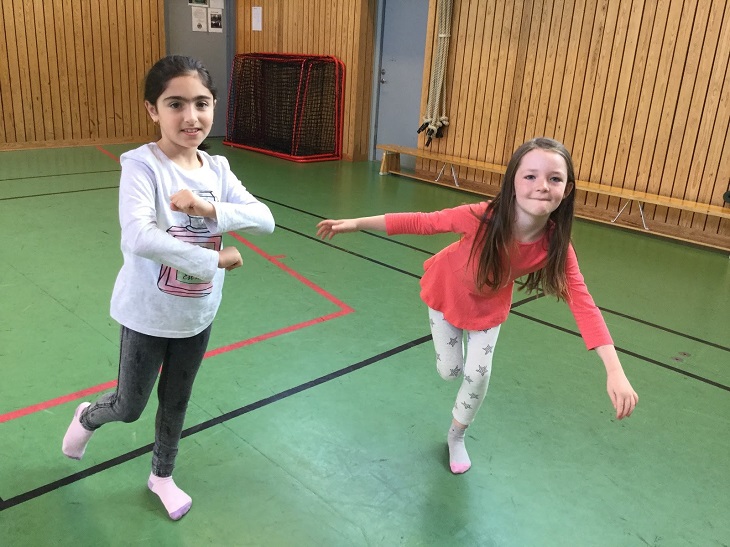Judith och Kavi står i en gymnastiksal och dansar. En av dem snurrar armarna framför sig och den andra står på ett ben med armana utåt.