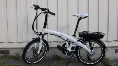 En elcykel står framför en vit vägg. Den har små hjul och en vit ram som är ganska bred. Under pakethållaren syns en liten svart låda.