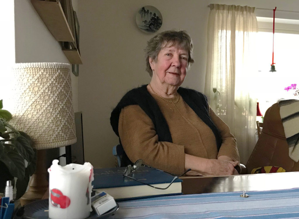 Dorothy Samuelsson har en svart väst över en brun tröja. Hon sitter vid ett bord i sitt vardagsrum.