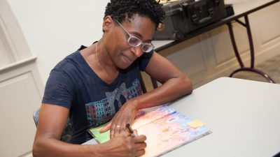 Jacqueline sitter vid ett bord. Hon skriver längst fram i en barnbok. Jacqueline har mörkt kort hår, glasögon och en mörkblå t-shirt.