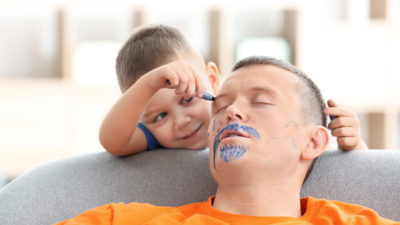En person ligger och sover i en fåtölj. Två barn har smugit upp bakom personen och målar en mustasch och skägg utan att han märker något.