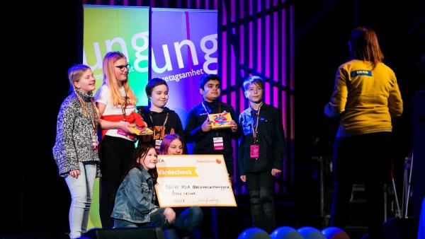 Sex elever står på scen. Några håller i en stor värdecheck och några håller i varsin ask med choklad.