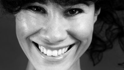 Miriam ler stort mot kameran. Hon har mörk lockigt hår som faller ner i pannan. Ögonen glittrar.